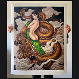 artist holds a print of "dragon" - a woman with large eyes and long dark hair stands with her back to the viewer, a golden dragon is curled around her and they are both surrounded by soft gray clouds